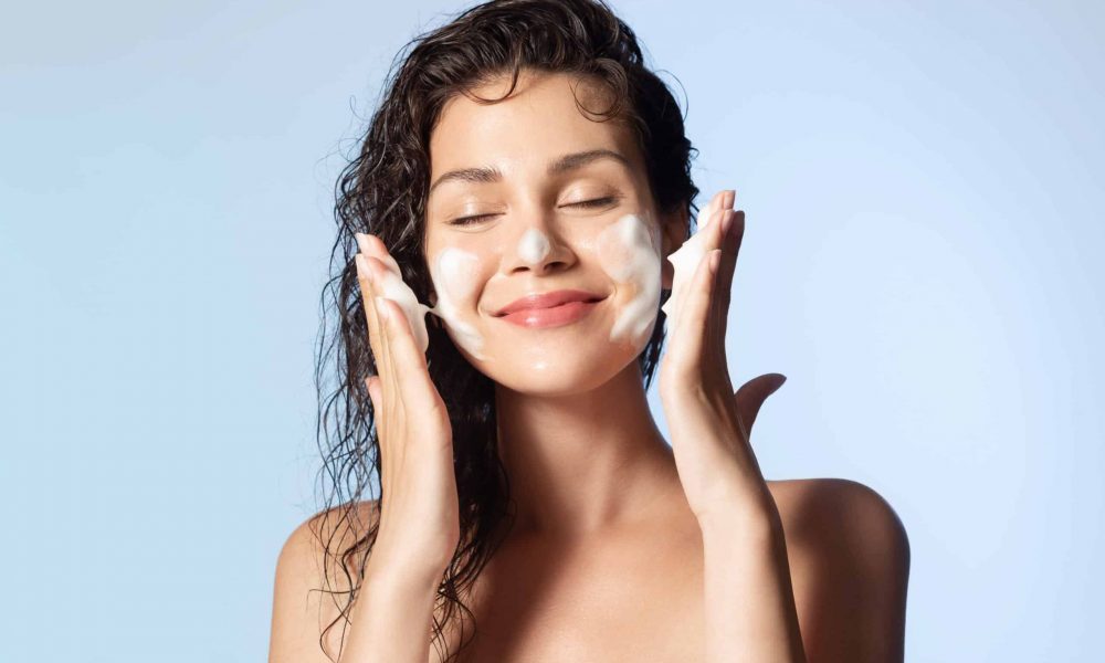 Smiling young woman washing foam face by natural foamy gel. Satisfied girl with bare shoulders applying cleansing beauty product on cheeks closed eyes. Personal hygiene, skincare daily routine.