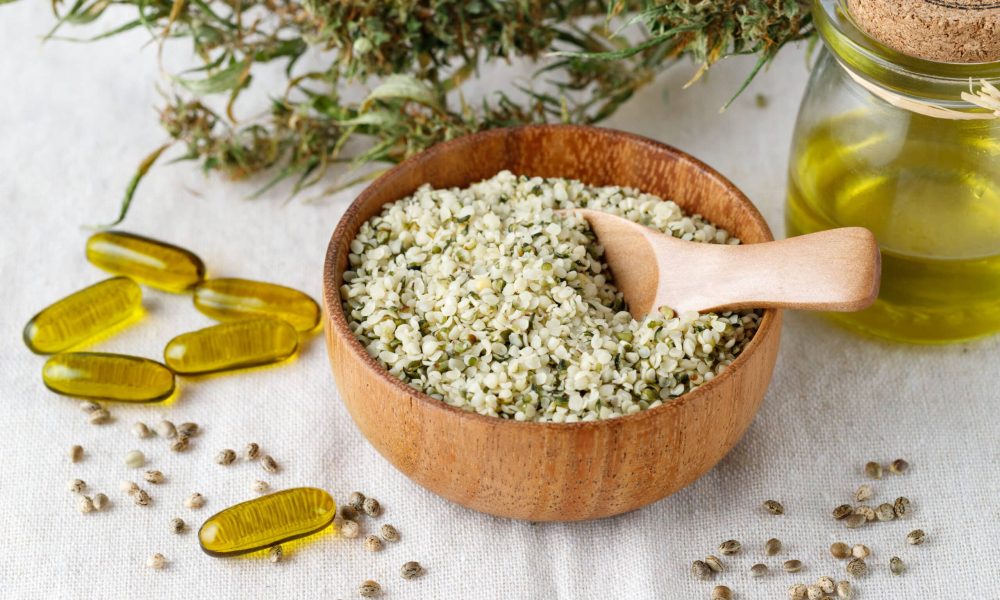 Shelled hemp seeds and oil as superfoods, supplement for eat with fiber and omega 3. Crushed cannabis seeds in wooden bowl with spoon  and cbd oil capsules on hemp fabric. Hemp products concept.